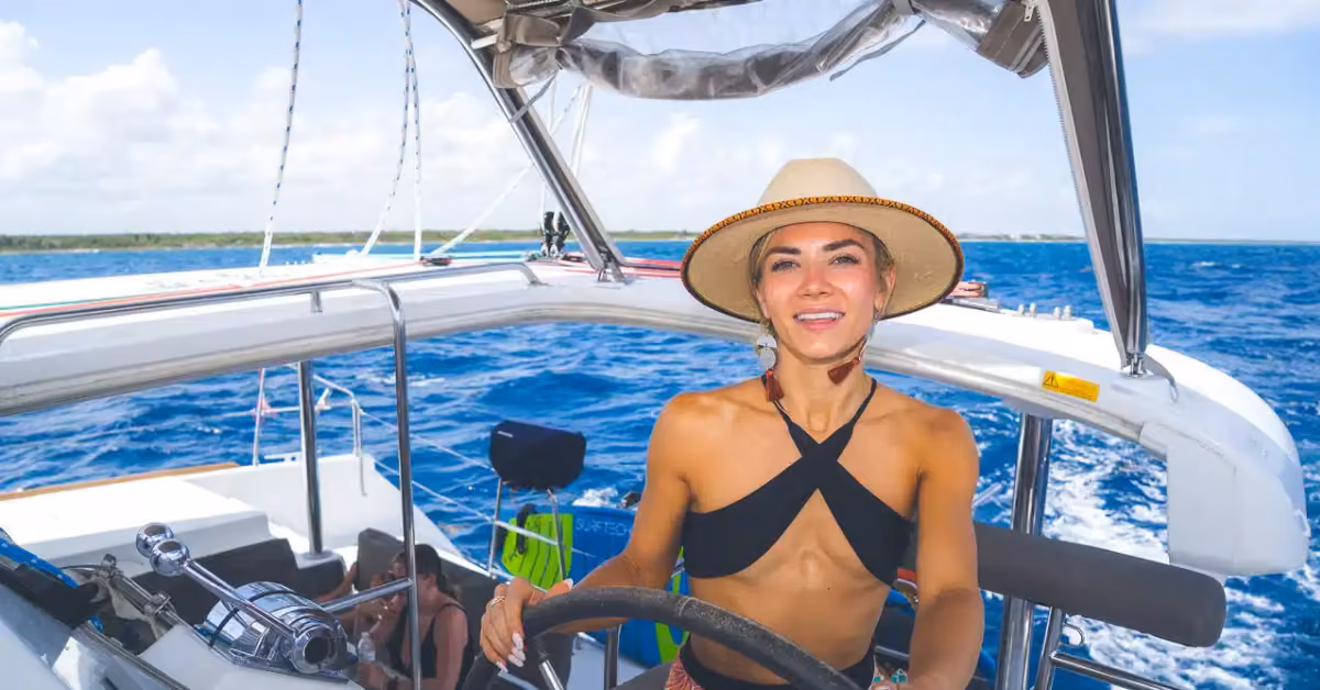 A smiling woman in a sunhat and bikini top steers a sailboat across blue water with distant land under a partly cloudy sky.