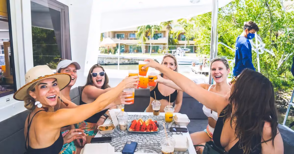 Six friends toast around a table on a cruise, smiling with food and drinks, lush greenery and sparkling water behind them.