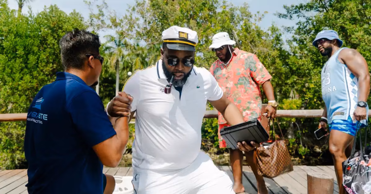 A man in white and a captain’s hat is helped off a boat, followed by two men, on a wooden dock surrounded by greenery.