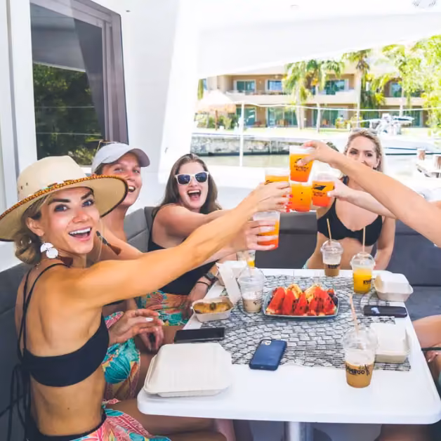 Five women toast with orange drinks around a table of food on a boat, smiling in the sun with sparkling water behind them.