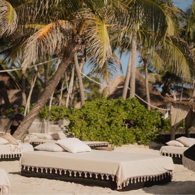 Beach beds with beige covers rest under palm trees on sand at Gitano Beach Club, with cabanas and lush bushes in the background.
