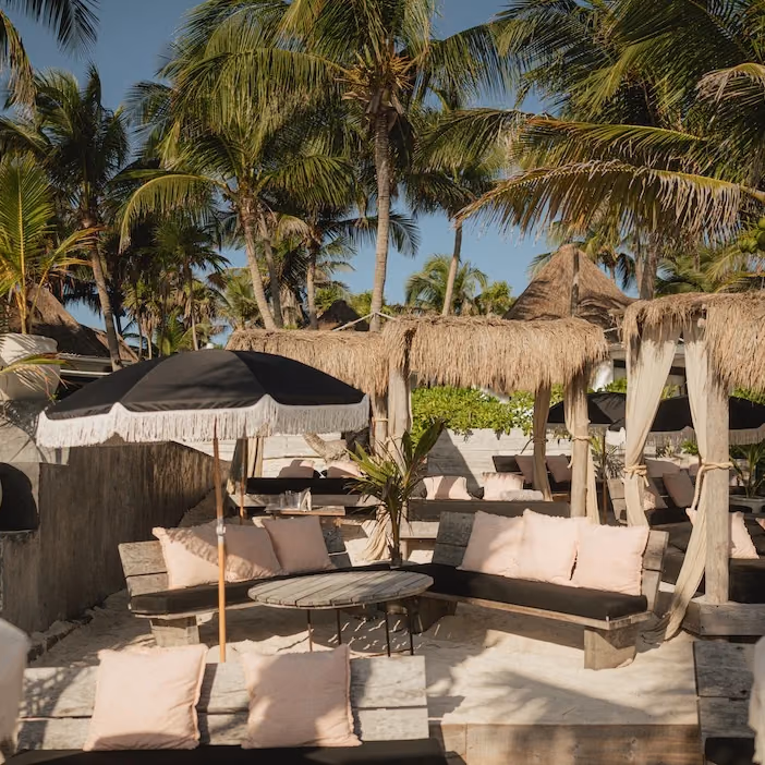 Beach Suite at Gitano Beach: black benches with pink cushions around a wooden table under a fringed black umbrella, thatched cabanas and palm trees behind.
