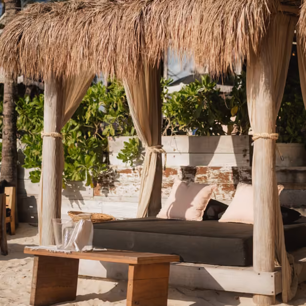 A thatched-roof cabana with sheer curtains and lounge seating sits on sand, surrounded by plants and wooden benches.