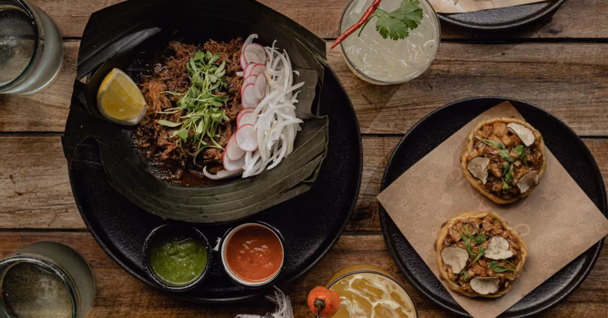 Mexican dishes and drinks on a wooden table: shredded meat with radishes, salsas, tostadas, and colorful garnished drinks.