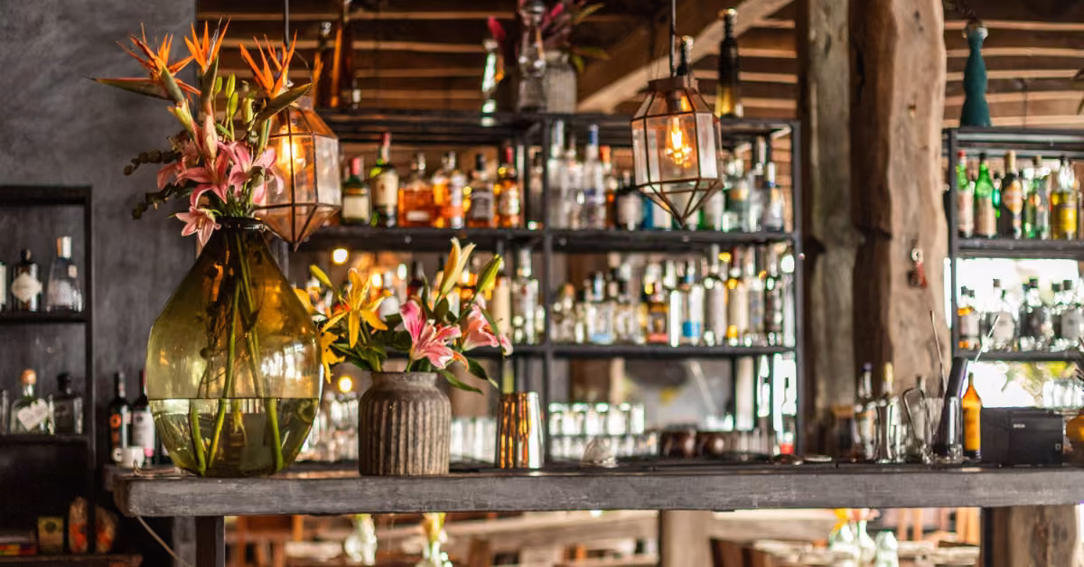 Cozy rustic bar at Gitano Beach Club with shelves of liquor, pendant lights above, and fresh flowers on the wooden counter.