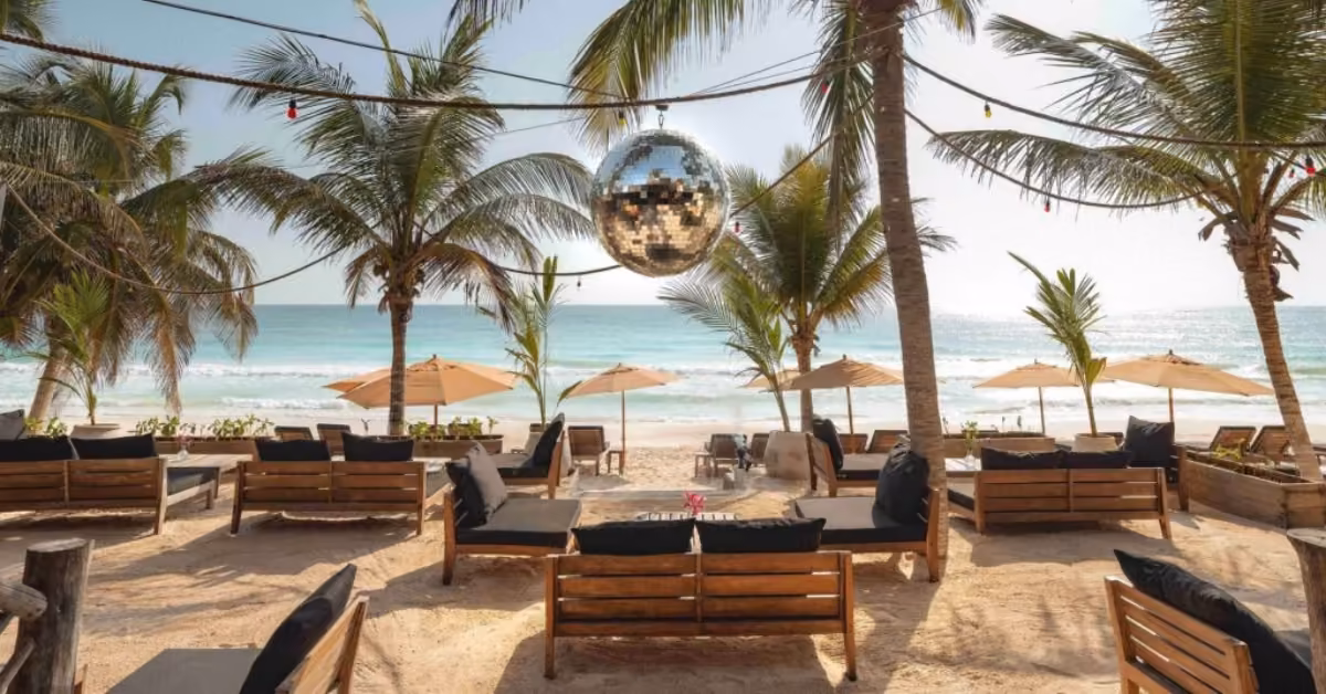 Gitano Beach Club lounge with wooden sofas, black cushions, disco ball, palm trees, umbrellas, and ocean under a sunny sky.
