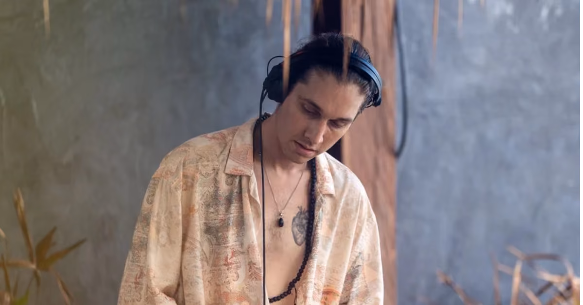 A person with long dark hair, headphones, and a patterned shirt stands indoors by a gray wall at Gitano Beach Club, looking focused.