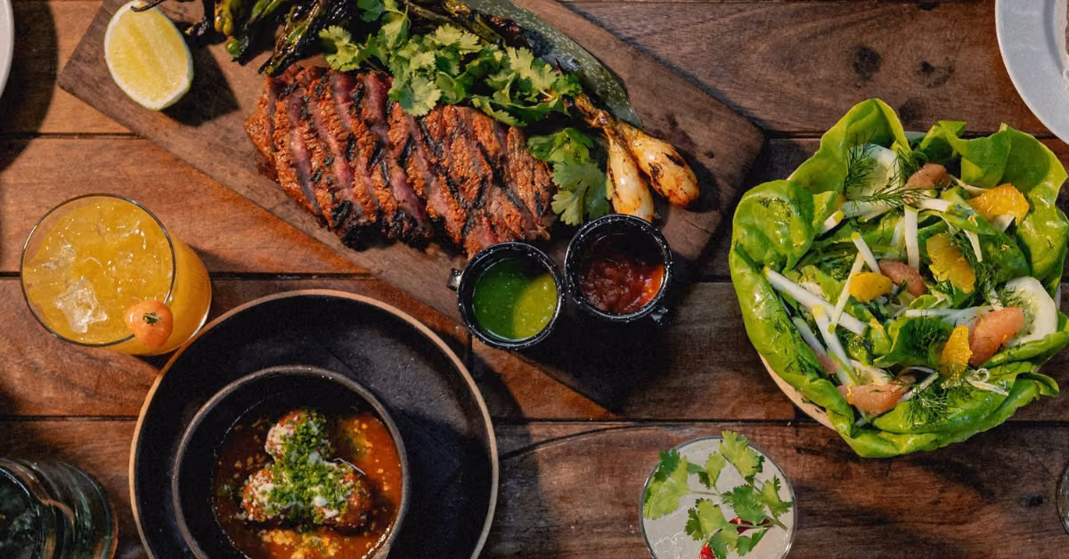 Grilled steak, salsa, roasted onions, citrus salad, meatballs, and citrus drinks on a rustic table at Gitano Beach Club.