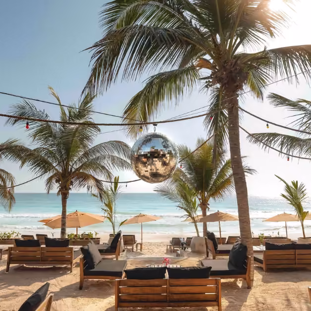 A shiny disco ball hangs between palm trees above a beach lounge with cushioned seats, umbrellas, and an ocean view.