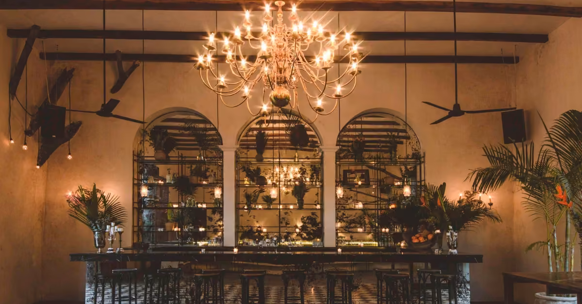 A dim, elegant bar with a large chandelier, arched mirrors, hanging plants, bar stools, and cozy warm ambient lighting.