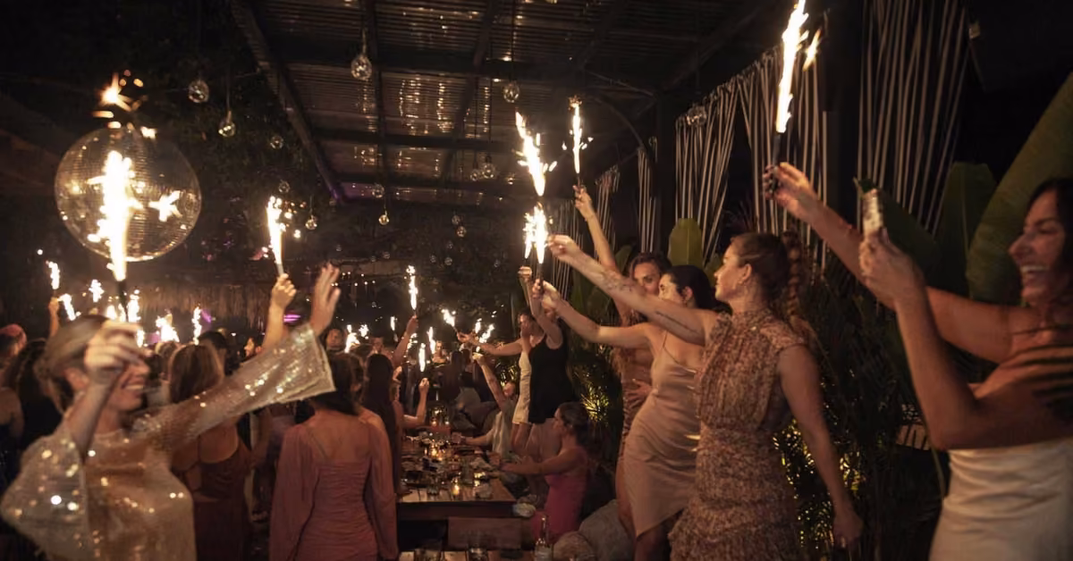 A joyful group celebrates with sparklers at a long table of food and drinks, surrounded by lush plants and festive decor.