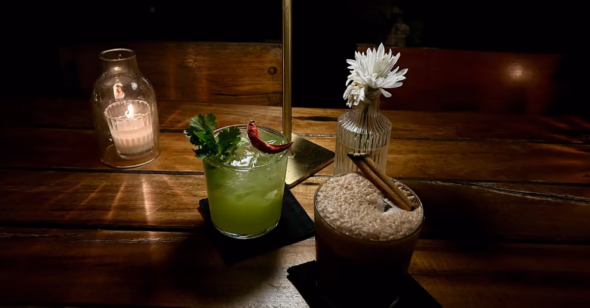 Two cocktails on a wooden table (one green with cilantro and chili, one brown with cinnamon) plus a lit candle and white flower.
