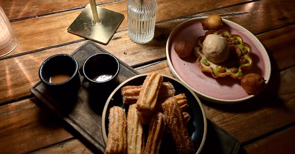 A table with churros, dipping sauces, waffles, ice cream, and pastries sits under warm lighting at Gitano Jungle Restaurant.