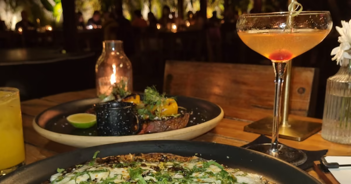 A warmly lit restaurant table with a cocktail, yellow drink, herb-garnished dish, vegetables, candle, and flower in a vase.