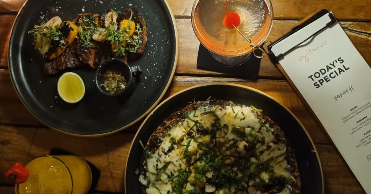 Overhead view of a wooden table at Gitano with two plates, two cocktails, a lime wedge, and a “Today’s Special” menu.