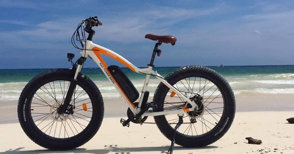 A white and orange electric fat-tire bike on a sandy beach, ocean waves and blue sky in the background.