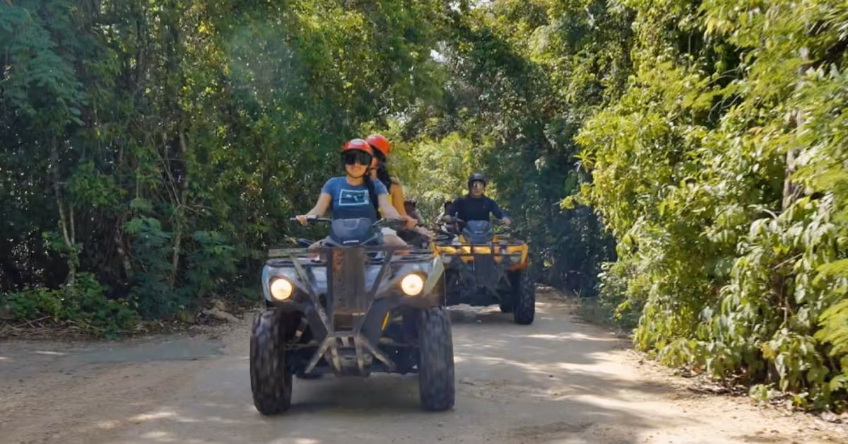 Three people ride ATVs down a forested dirt path; the lead wears a red helmet as sunlight filters through the trees.
