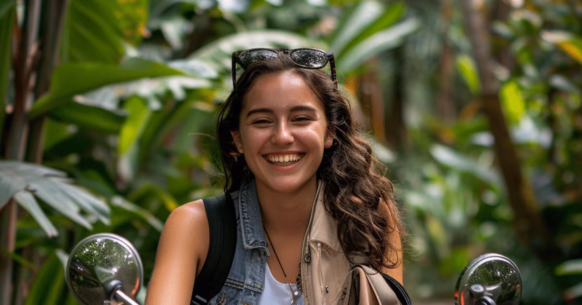 Smiling woman with long wavy hair sits on a scooter in Tulum, wearing sunglasses, denim vest, and backpack amid lush greenery.