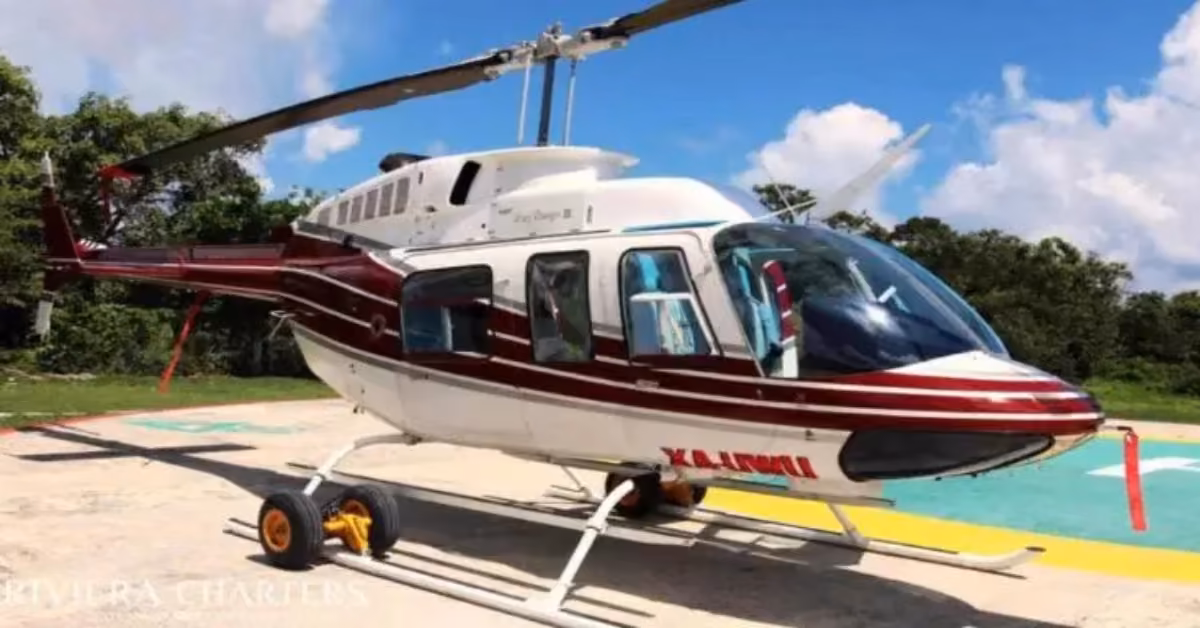 A white helicopter with red stripes, marked KANNI rests on a helipad amid greenery beneath a clear blue sky.