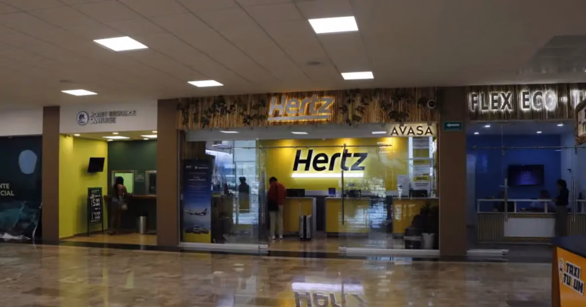 A Hertz rental car counter with yellow signage inside an airport, next to Flex Eco and Budget, people waiting at each.