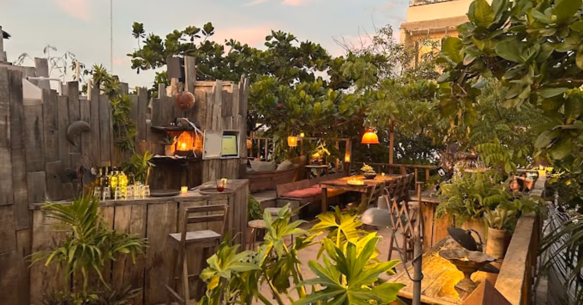 Cozy patio with rustic wood furniture, warm lights, lush plants, a small bar, and seating surrounded by trees at sunset.