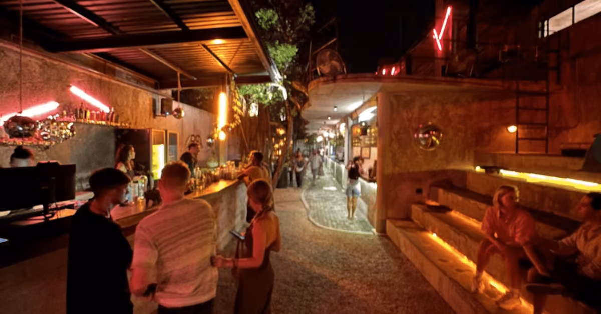 Nighttime outdoor bar with people socializing, some on benches, others at the bar; warm yellow lights and neon create a lively vibe.