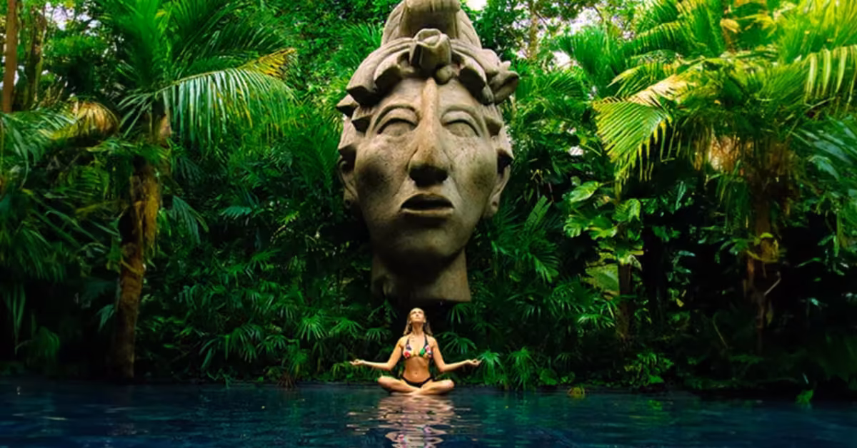 A woman in a bikini meditates cross-legged by a pool at Cenote Atik, lush jungle and a stone statue head behind her.