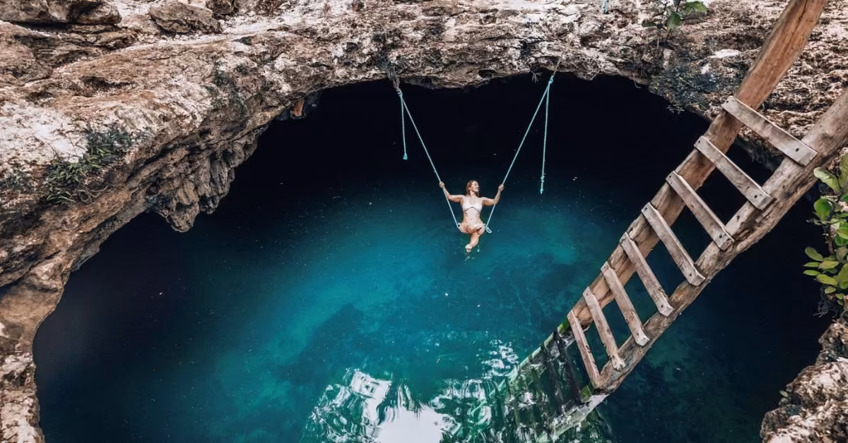 A person swings over clear blue waters at Cenote Calavera, surrounded by rocks with a wooden ladder leading into the cenote.
