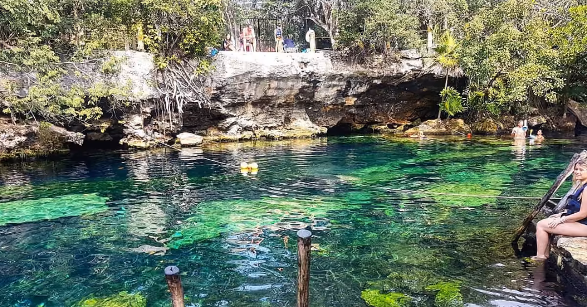 A turquoise cenote surrounded by rocks and greenery, with people swimming and relaxing at the water’s edge.