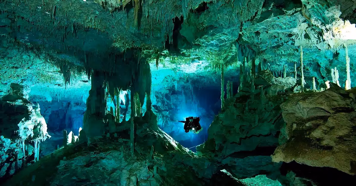 A diver swims through glowing caves in Cenote Dos Ojos, surrounded by turquoise water, stalactites, and rocky shapes.