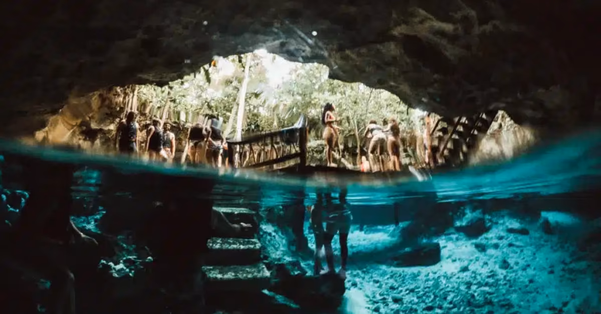 People stand on a wooden bridge above Gran Cenote’s clear blue water, with rocks and swimmers visible below in the cave.