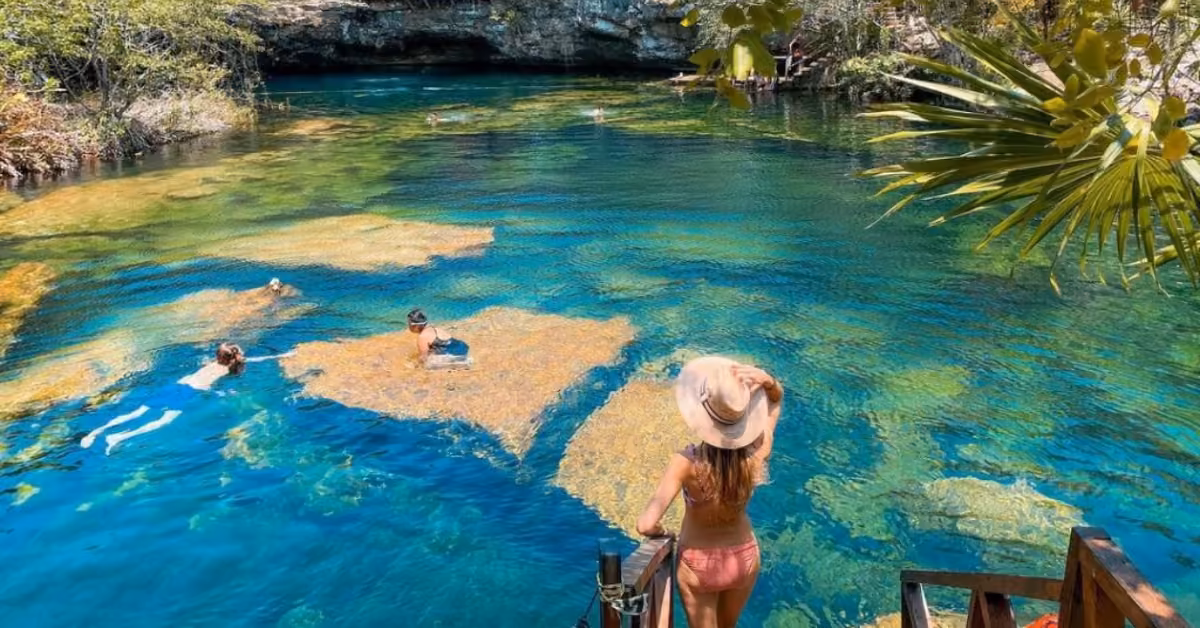 A woman in a hat stands on wooden steps by clear blue water, watching swimmers in a natural pool surrounded by lush greenery.