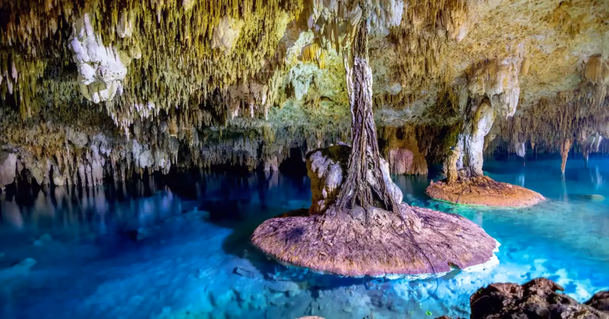 An underground cave with clear blue water, stalactites above, rocky formations, and gentle natural and artificial lighting.