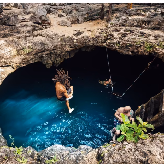 A person with dreadlocks jumps into a deep blue cenote as others swim and relax nearby, surrounded by rocks and lush greenery.