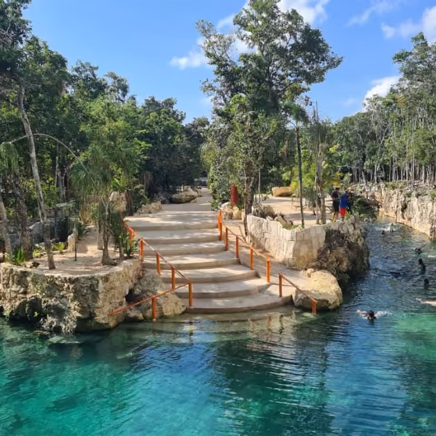 A stone staircase with railings leads to a lush, tree-filled area bordered by turquoise water where people swim and relax in the sun.