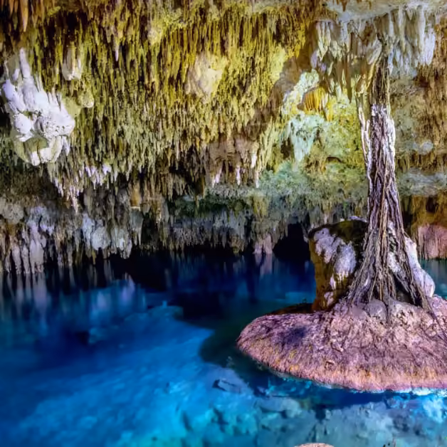 A cave with hanging stalactites, clear blue water on the floor, and a large central stalagmite on a rocky island.