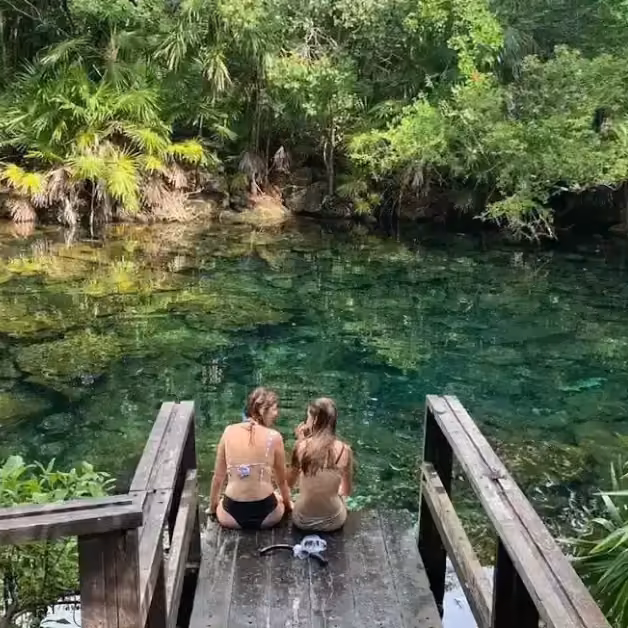 Two people sit on a wooden dock over a clear green-blue pool, surrounded by lush trees in a peaceful, tropical setting.