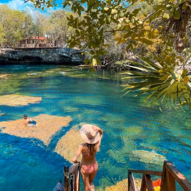 A woman in a hat and pink swimsuit stands on wooden steps, overlooking a sunlit cenote with clear blue water and swimmers below.