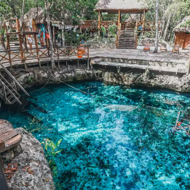 A clear blue cenote with wooden walkways, thatched huts, and lush greenery, sunlight shimmering on the water.