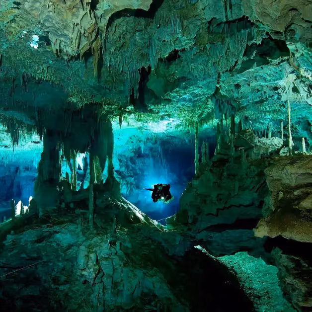 A scuba diver swims through an underwater cave with striking stalactites, lit by soft blue-green light.