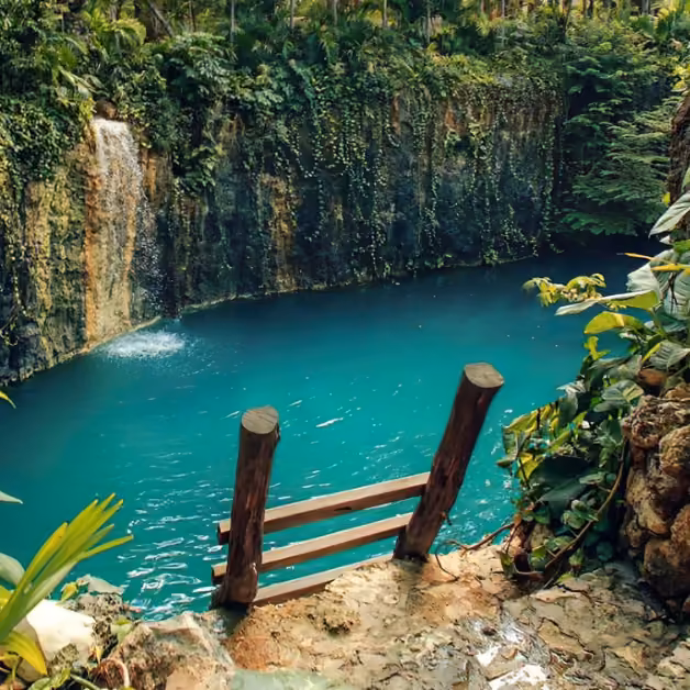 A wooden railing overlooks a bright blue cenote with lush plants and vines, and a small waterfall spilling into the water below.