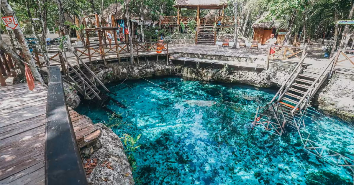 Blue cenote with wooden decks and ladders amid trees, sunlight filtering through leaves. Small huts and a swimmer nearby.