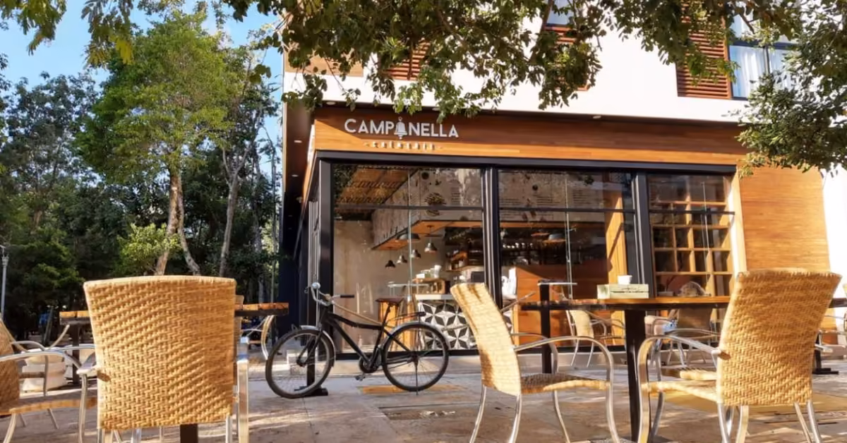 Outdoor café seating with wicker chairs and tables at Campanella. A bike is parked nearby under tree shade with dappled sunlight.