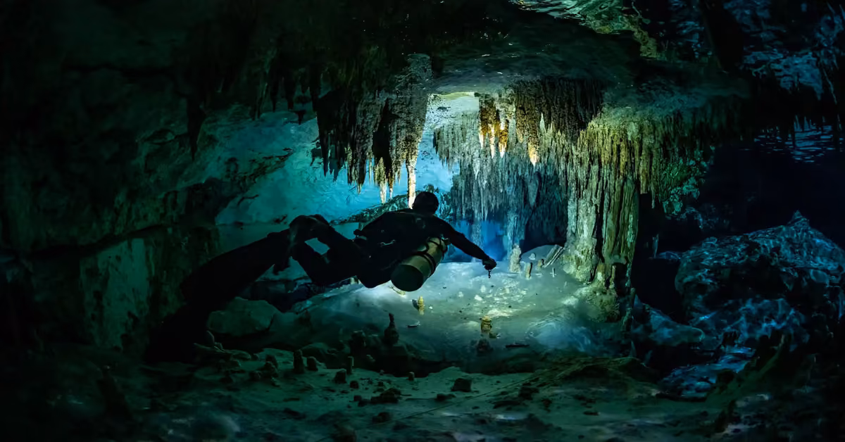 A scuba diver swims through an underwater cave lit by blue-green light, with stalactites hanging above and a sense of adventure.