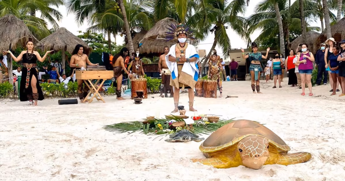 A beach ceremony with dancers, traditional performer, drums, onlookers, turtle sculpture, palm trees, and nearby huts.