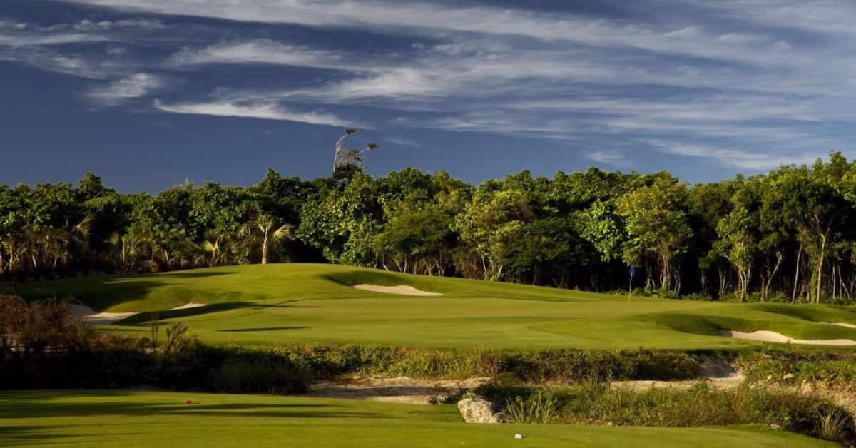 Lush golf course with green fairways, sand bunkers, and dense trees beneath a blue sky dotted with wispy clouds near Tulum.