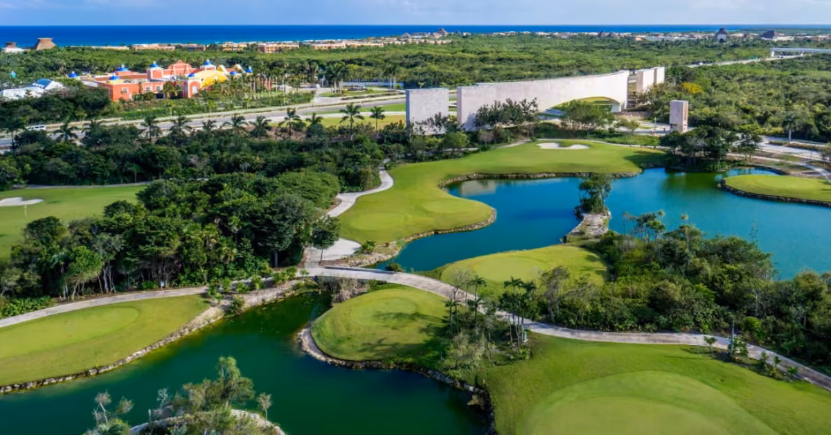 Aerial view of a lush golf course with ponds, winding paths, green landscape, and colorful buildings under a bright blue sky.