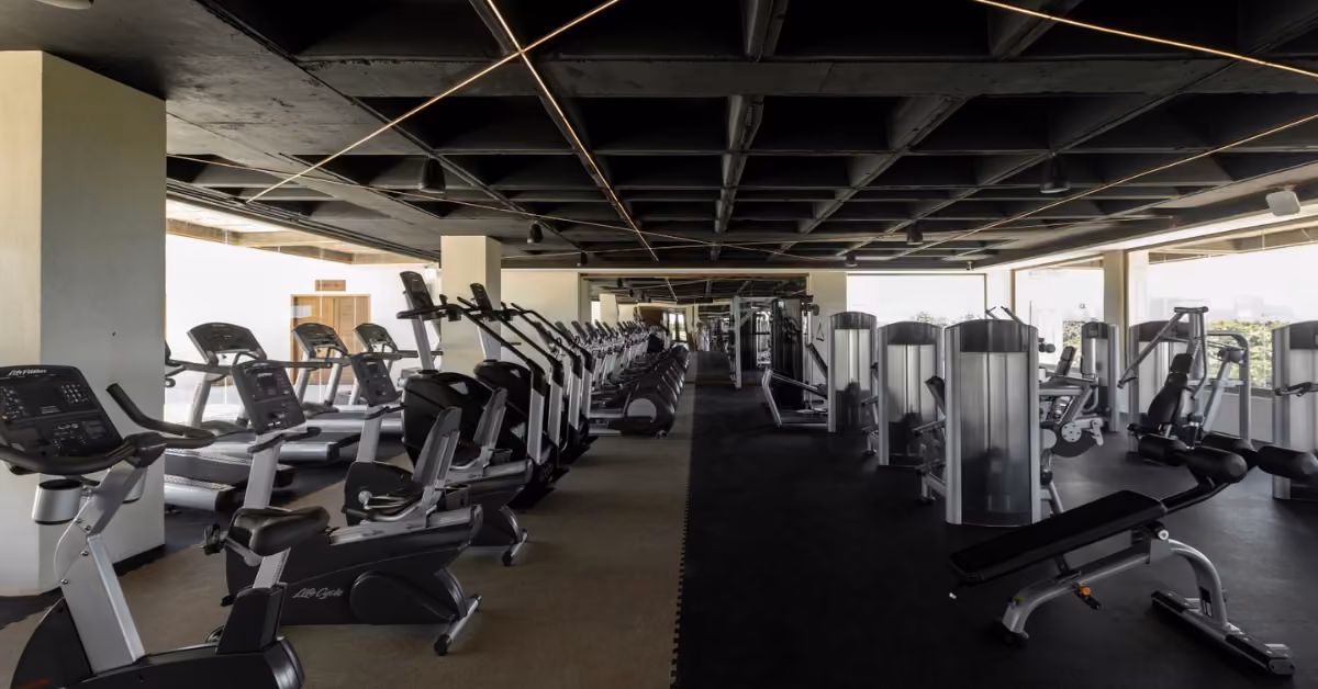 Modern gym with exercise bikes, treadmills, and weight machines under geometric lights; large windows bring in natural light.