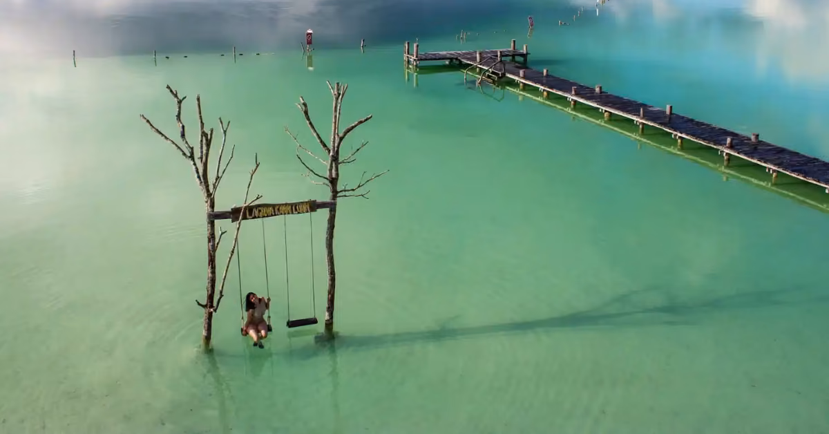 A person relaxes on a swing between two trees in clear turquoise water beside a wooden pier leading into the Tulum lagoon.