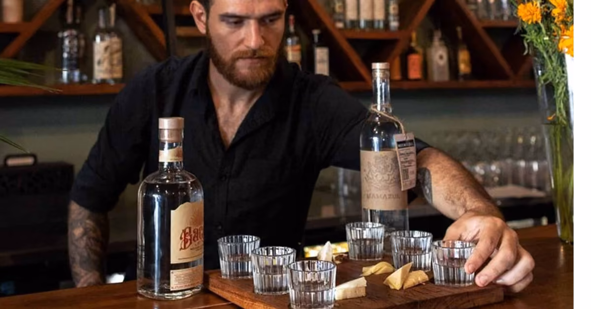 A bartender sets glasses, lime wedges on a tray, and bottles on the bar in Tulum, with liquor shelves behind.